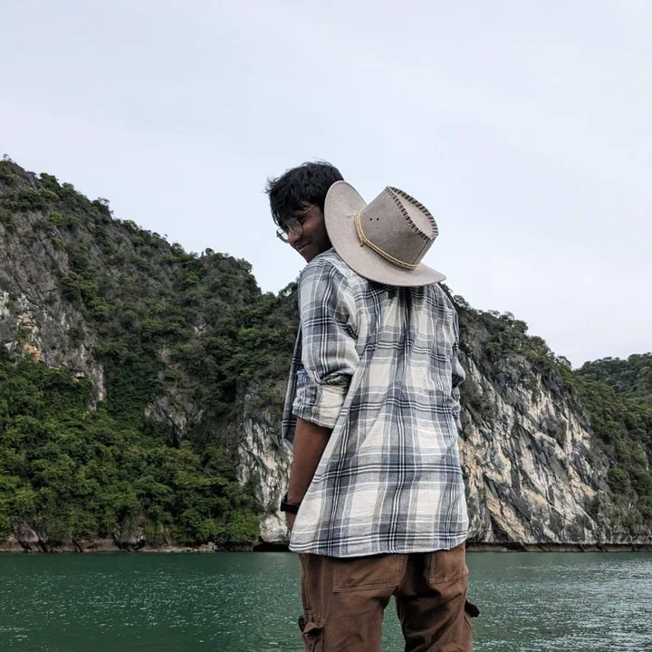 Abir in Vietnam — plaid shirt and travel hat, limestone cliffs of Ha Long Bay behind.